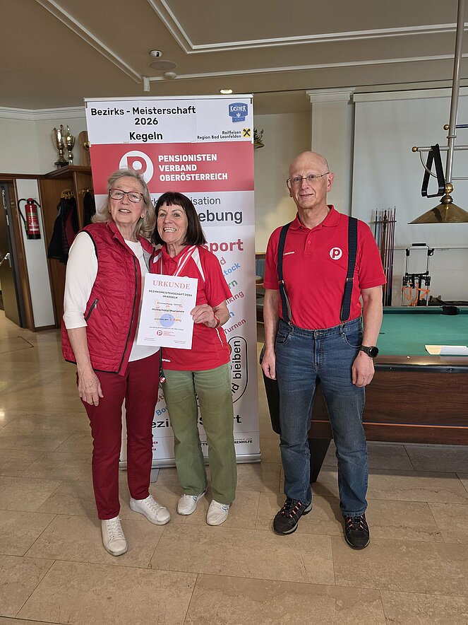 Zwei ältere Frauen eine blond die andere dunkel, diese hält Urkunde in der Hand und ein älterer Mann ohne Haare 