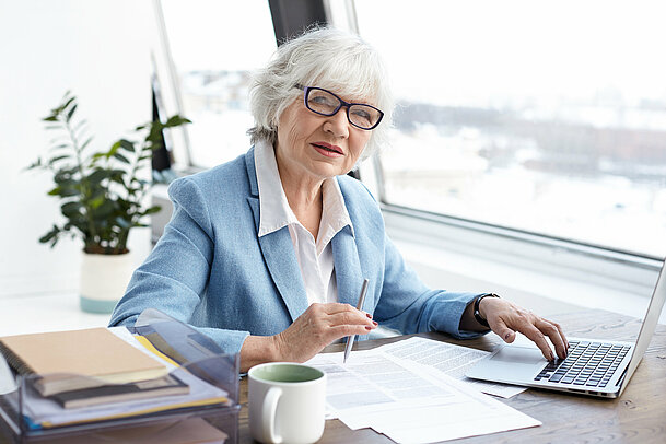 Attractive serious female chief executive officer of mature age sitting at her office with laptop, keyboarding and signing papers on desk, having confident look. People, aging, job and career concept Attractive serious female chief executive officer of mature age sitting at her office with laptop, keyboarding and signing papers on desk, having confident look. People, aging, job and career concept