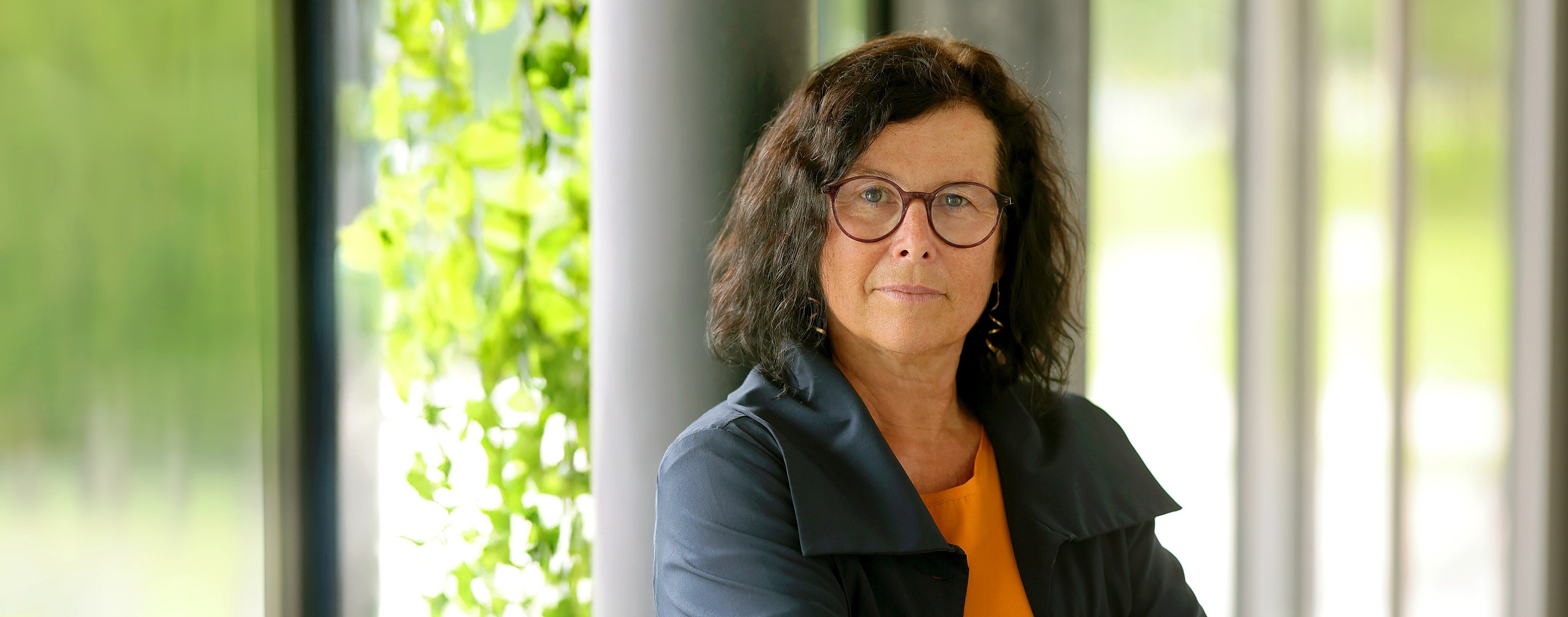 Frau mit Brille und dunklem Haar steht vor einem Fenster mit grünen Pflanzen