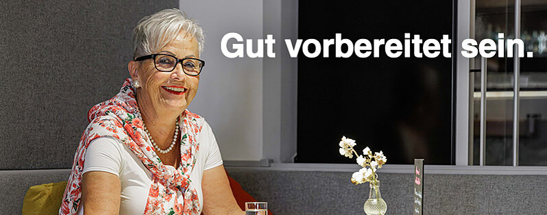 Lachende ältere Frau mit Brille und Blumen-Tuch sitzt in einem Büro.